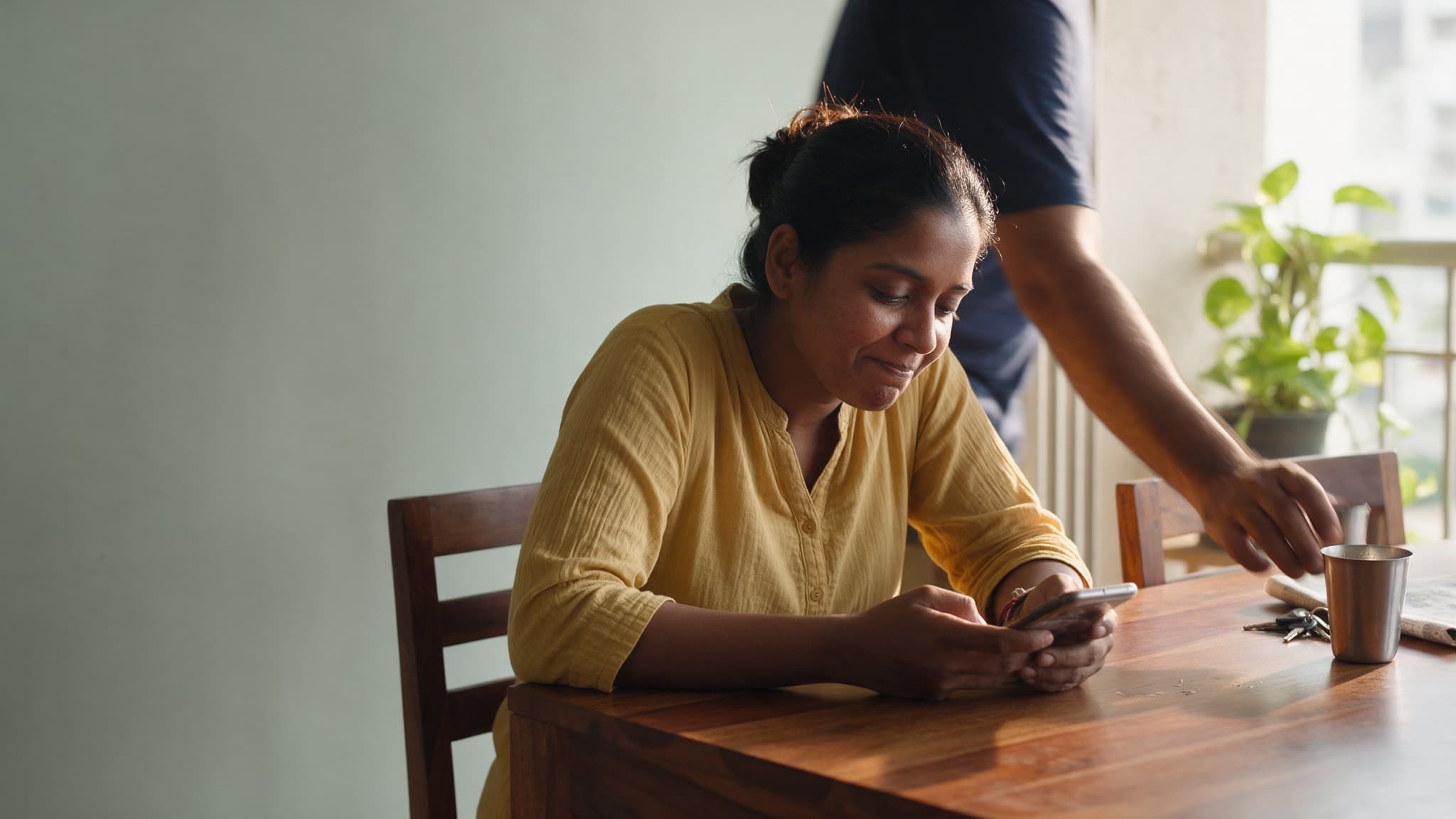 An Indian woman writing a message on her phone in morning light while her husband sets down a glass of chai