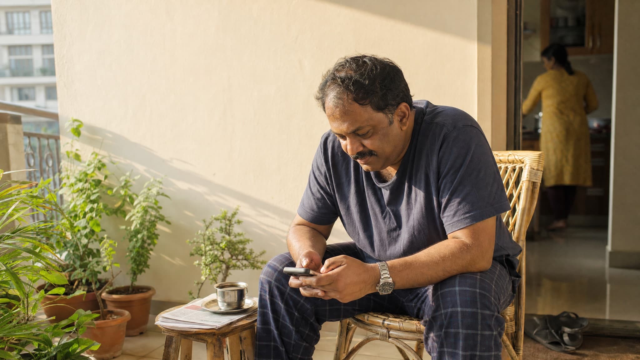 An Indian man on his balcony in early morning light, typing a long message on his phone while his wife is in the kitchen behind him