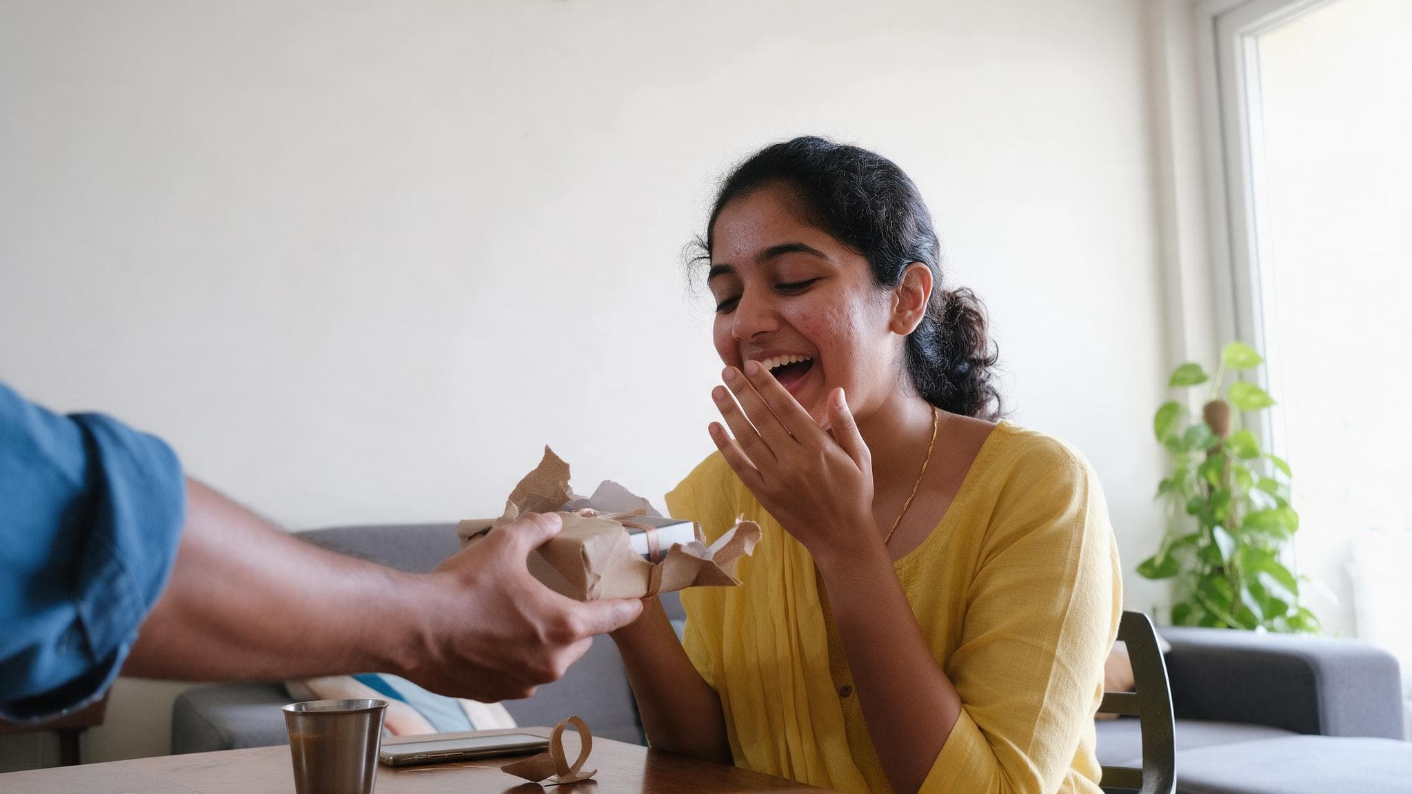 A young woman laughing as she opens a small wrapped birthday gift on a sofa in a bright flat, her boyfriend’s hand holding the box