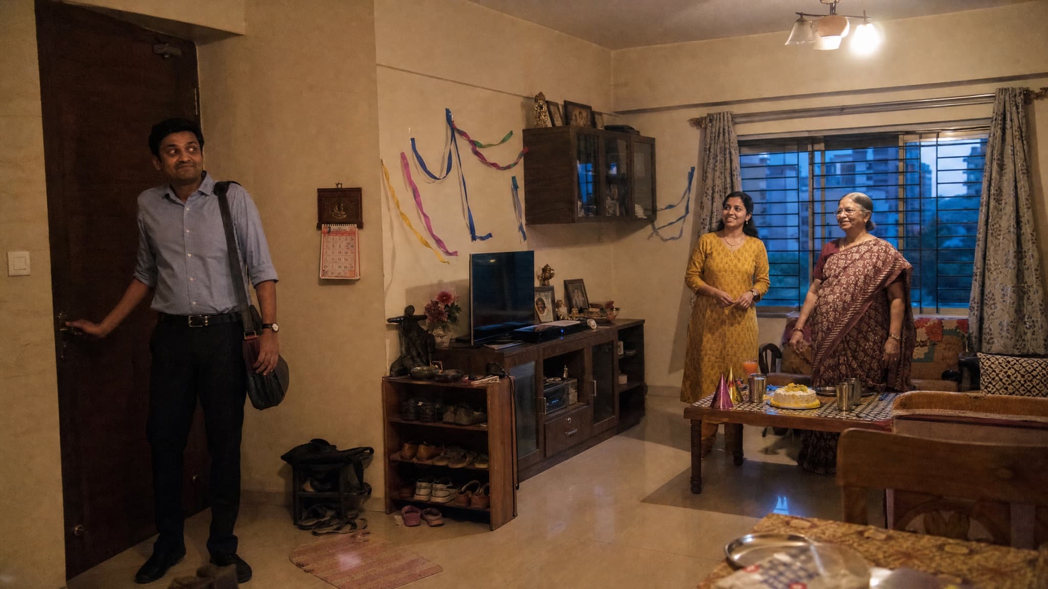 An Indian man in his thirties stopping in a doorway, surprised, as his wife and mother wait in the living room with a small bakery cake