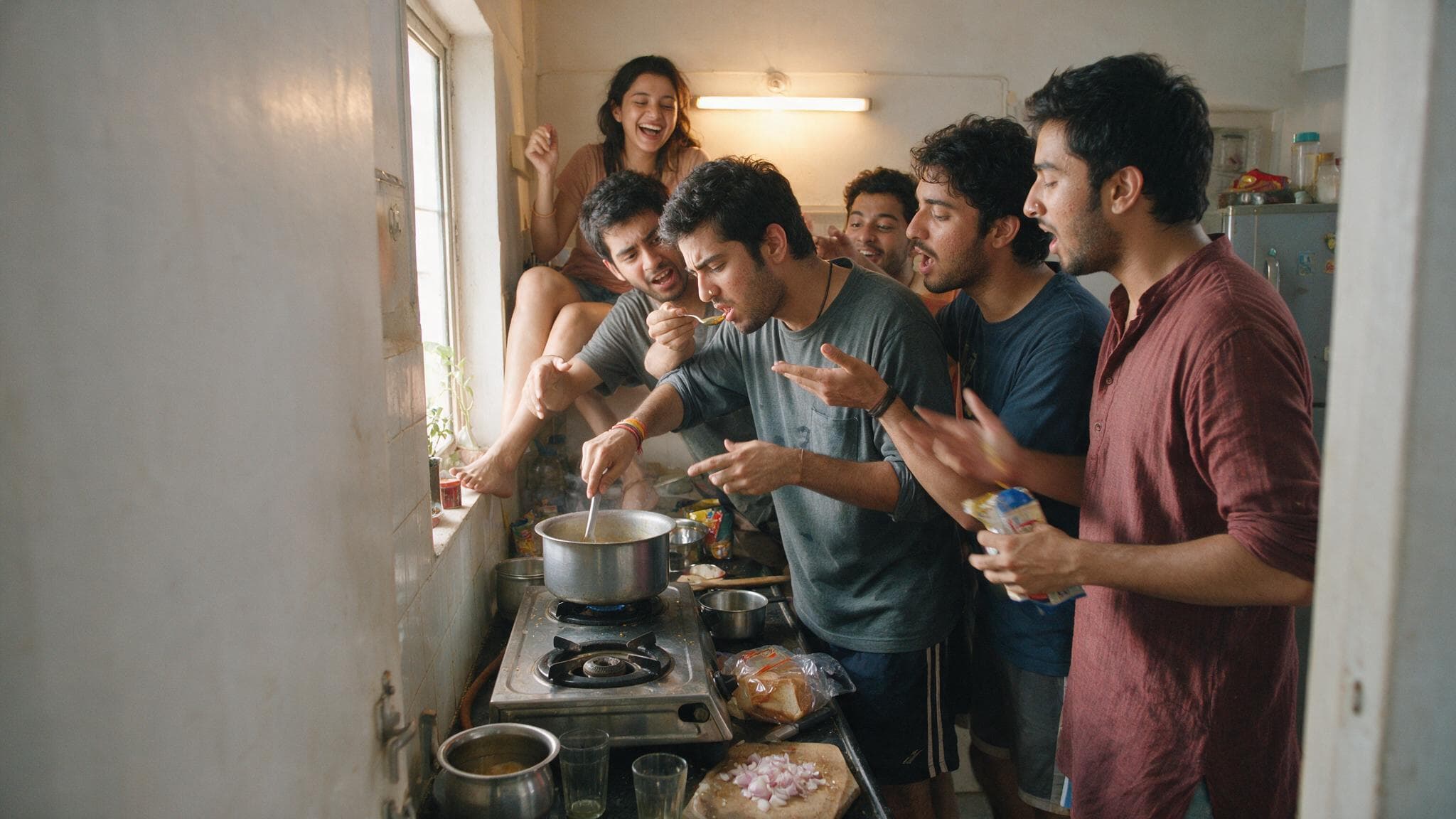 Six friends crowded into a small flat kitchen, one tasting from a pot while the others argue about it